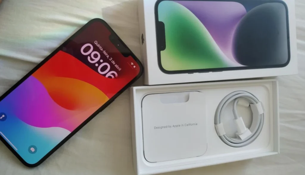 iPhone 14 Plus preto com tela ligada exibindo o relógio e data, posicionado ao lado da caixa aberta, revelando o cabo Lightning e o manual com a inscrição “Designed by Apple in California”.